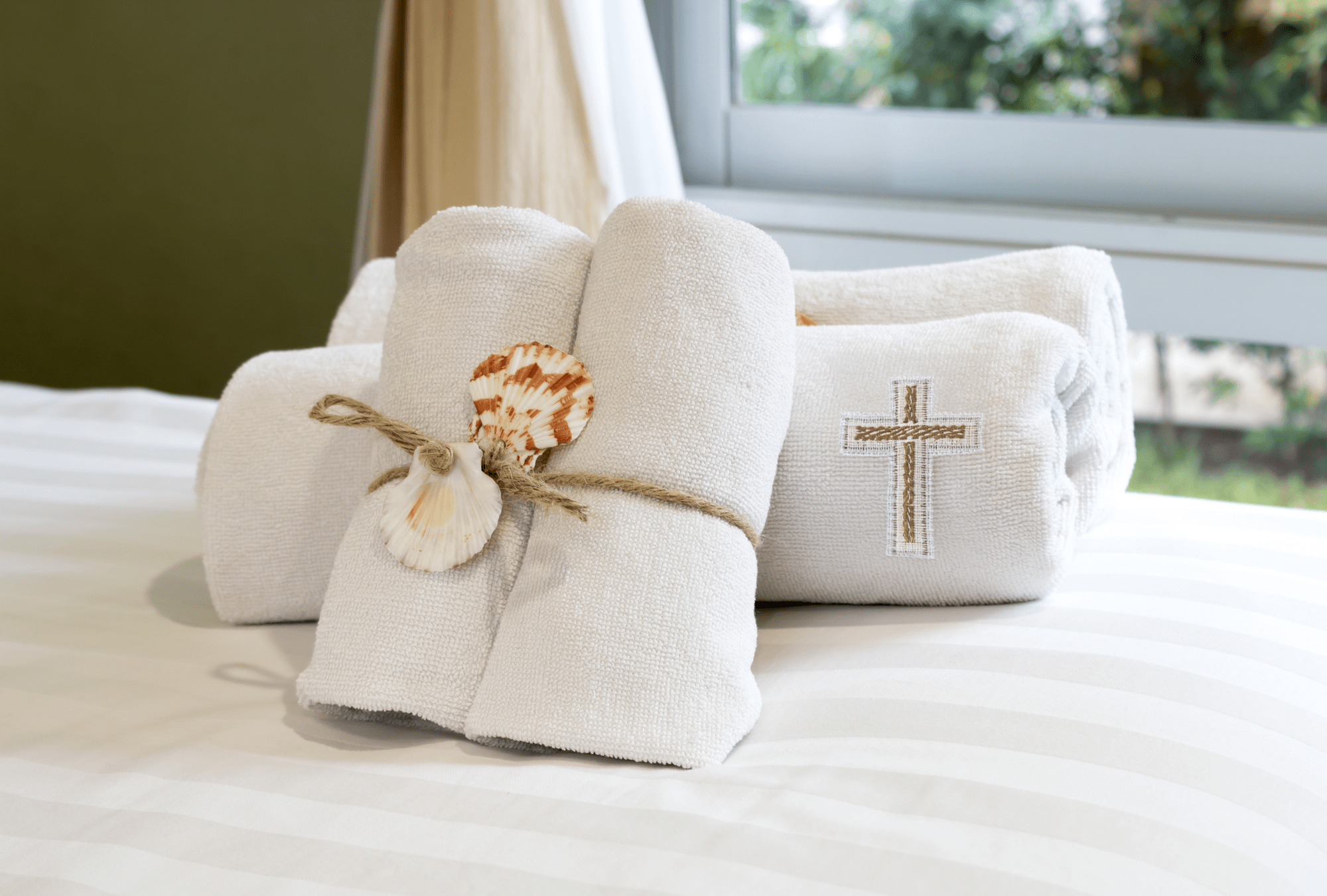 Soft, white towels neatly rolled and tied with twine, adorned with a seashell and a stitched cross