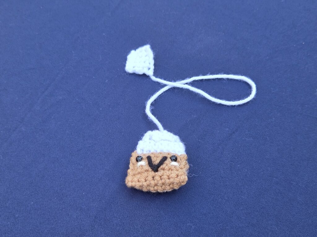 Crochet bookmark looks like a smiling tea bag. Brown and white with cute smiley face and safety eyes.