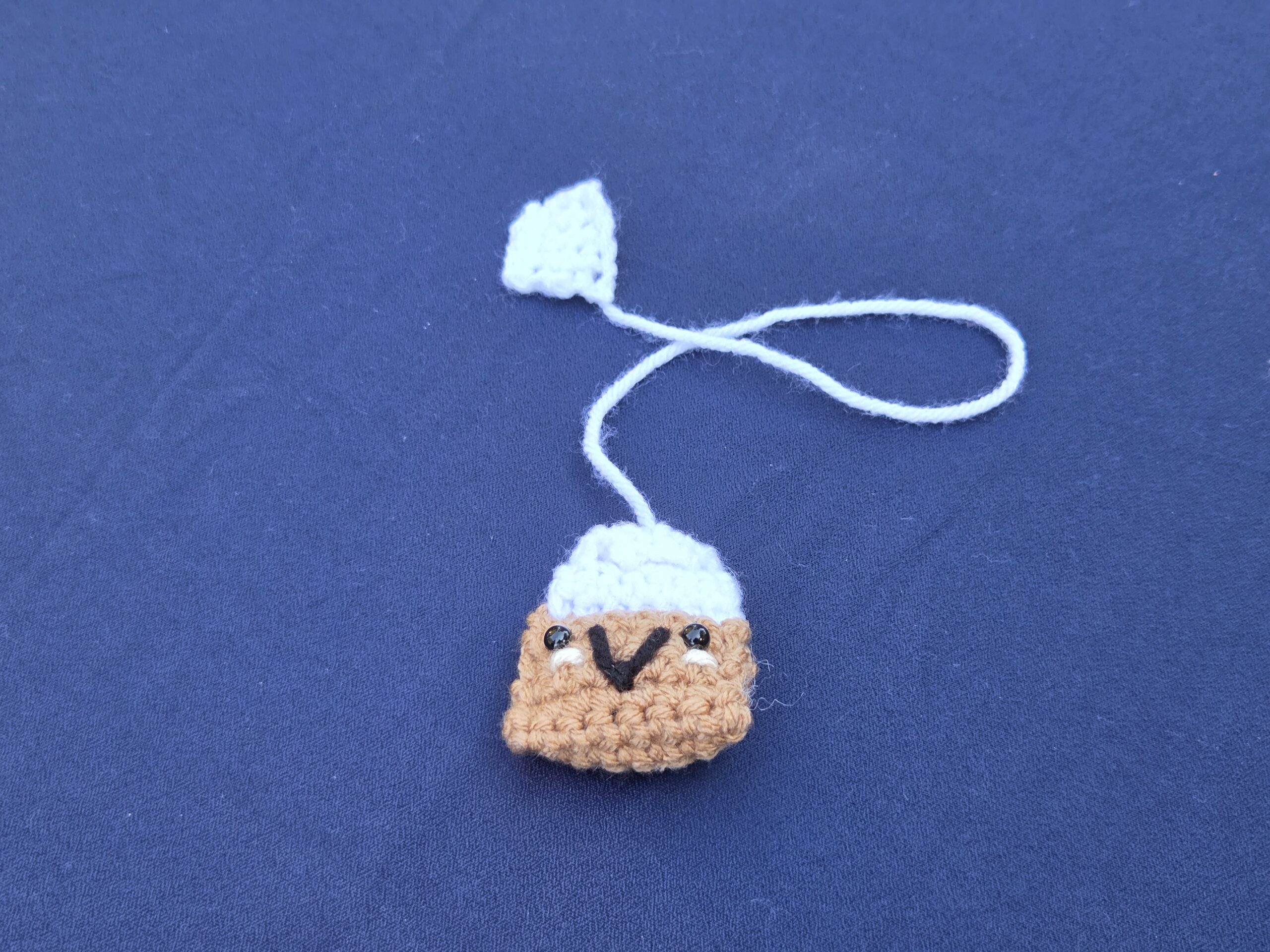 Crochet bookmark looks like a smiling tea bag. Brown and white with cute smiley face and safety eyes.