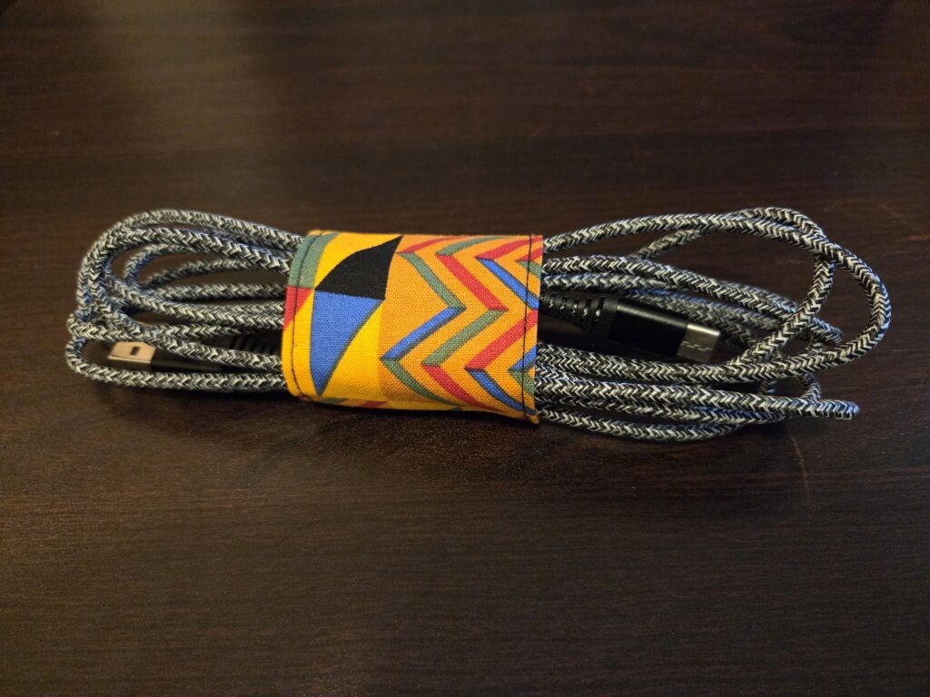 Wrap for keeping electronics cords. Jeweltones: deep gold with black, blue, red, and green geometric designs. Black Velcro enclosure. Sized for cell, tablet, or laptop cords. Cord shown not included.