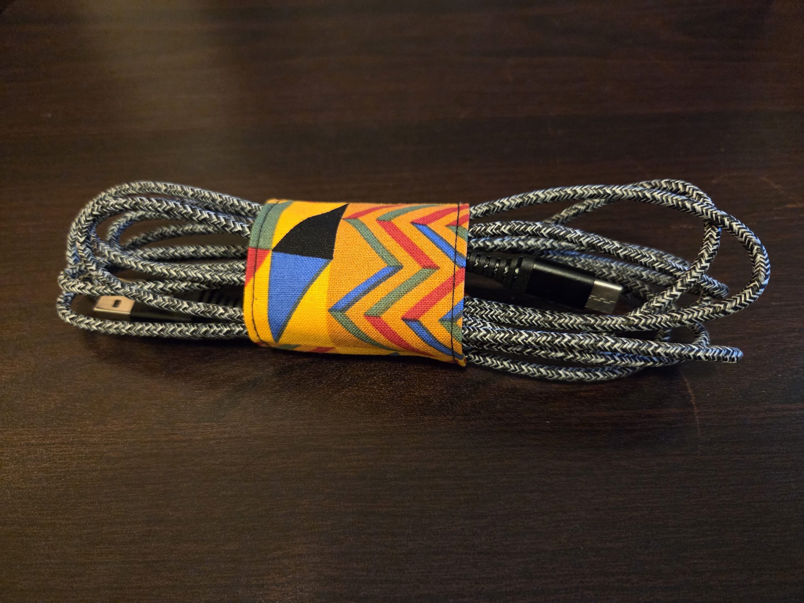 Wrap for keeping electronics cords. Jeweltones: deep gold with black, blue, red, and green geometric designs. Black Velcro enclosure. Sized for cell, tablet, or laptop cords. Cord shown not included.