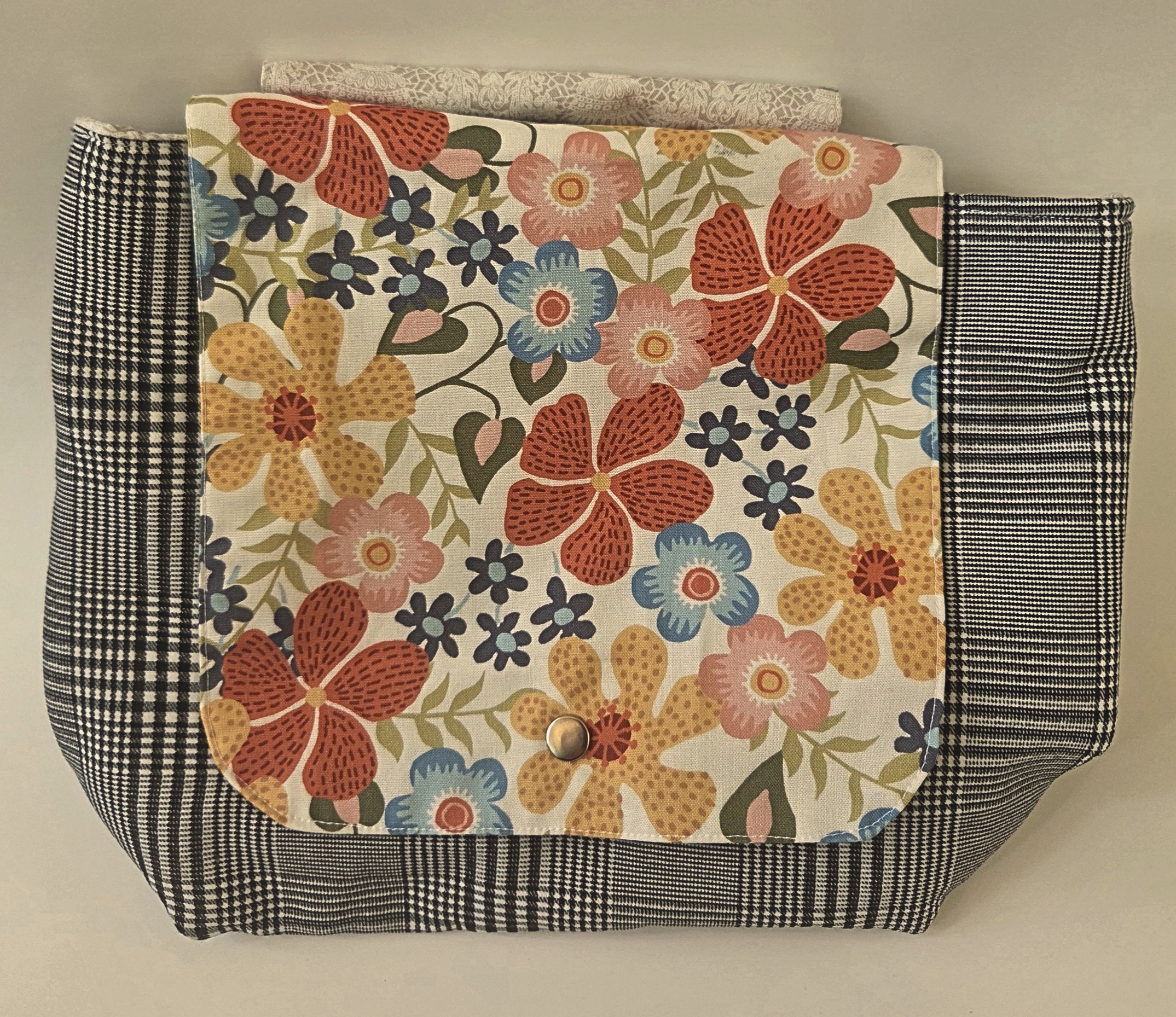 Front of messenger bag features flap with floral pattern in neutral colors. Bag body black and white plaid. Metal snap closure. Attaches to stroller, bike, walker, etc. with hook-and-loop tape strap (Velcro).