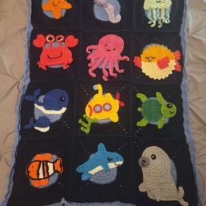 God's Sea Creatures crochet blanket features 12 crochet blocks, each with a marine animal: starfish, seahorse, crab, octopus, whale mom with baby, fish, submarine, shark, seal, turtle, pufferfish, and jellyfish. Dark blue background, with a line of light blue around the edges.