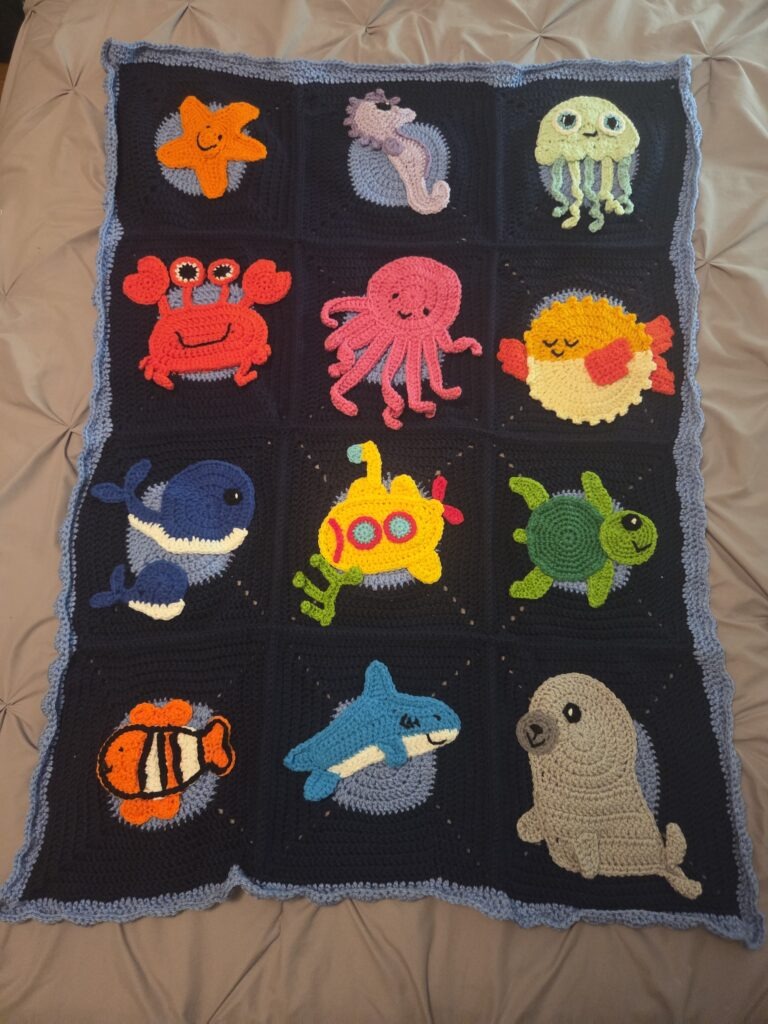 God's Sea Creatures crochet blanket features 12 crochet blocks, each with a marine animal: starfish, seahorse, crab, octopus, whale mom with baby, fish, submarine, shark, seal, turtle, pufferfish, and jellyfish. Dark blue background, with a line of light blue around the edges.